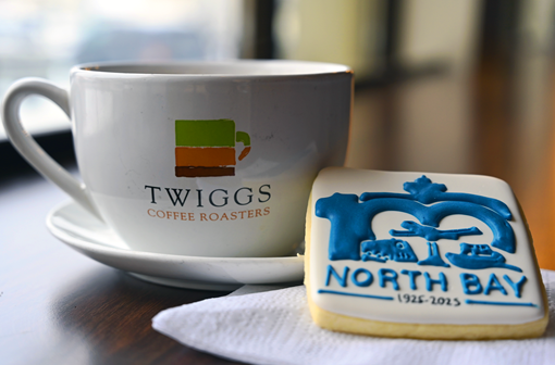 Twiggs branded white cup and saucer, next to it a square sugar cookie with white icing and North Bay Centennial logo in blue icing.